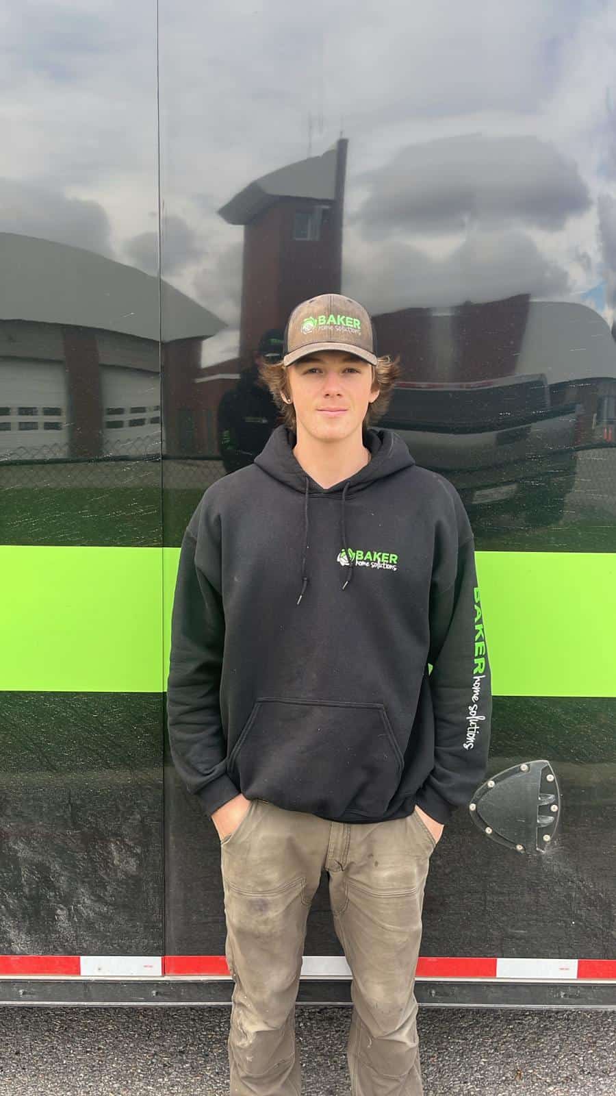Youthful worker in black Baker Home Solutions hoodie standing outdoors near a black and green vehicle.
