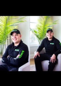 Roofing specialist wearing Baker Home Solutions apparel, sitting confidently indoors with lush green plant backdrop.