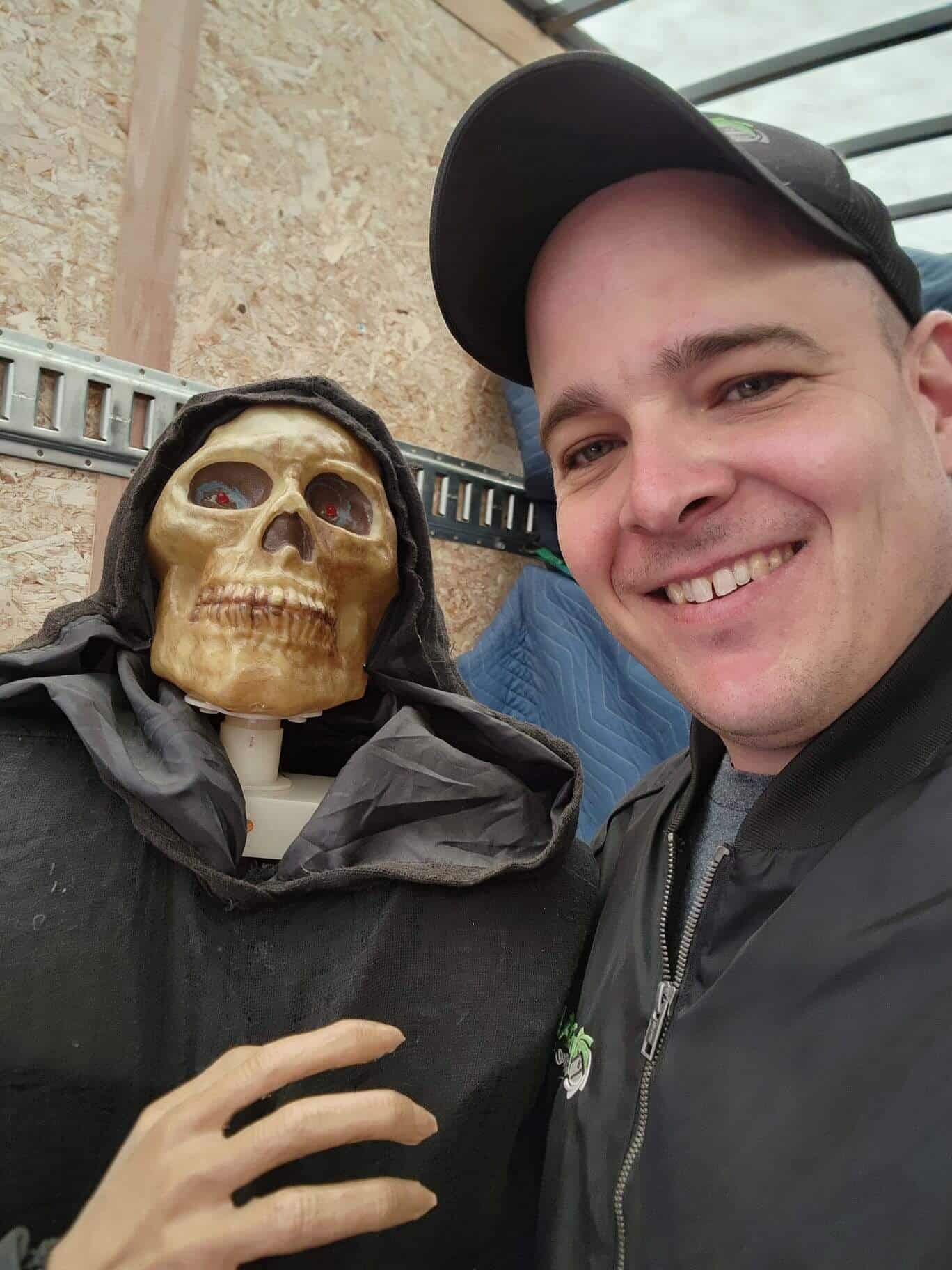 Skull mannequin with a black hood next to smiling man in a black jacket and cap.