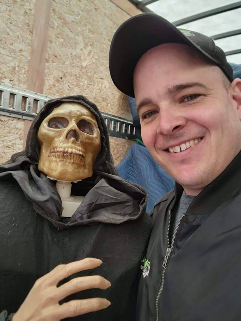 Skull mannequin with a black hood and a smiling man in a black jacket, outdoor setting.